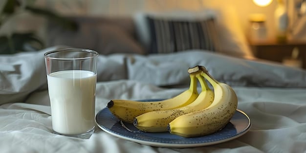 A glass of warm milk and a banana on a nightstand next to a bed.