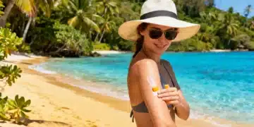Pessoa aplicando protetor solar em uma praia ensolarada, com chapéu e óculos de sol, simbolizando a preparação para o verão de 2026 e a proteção da pele.